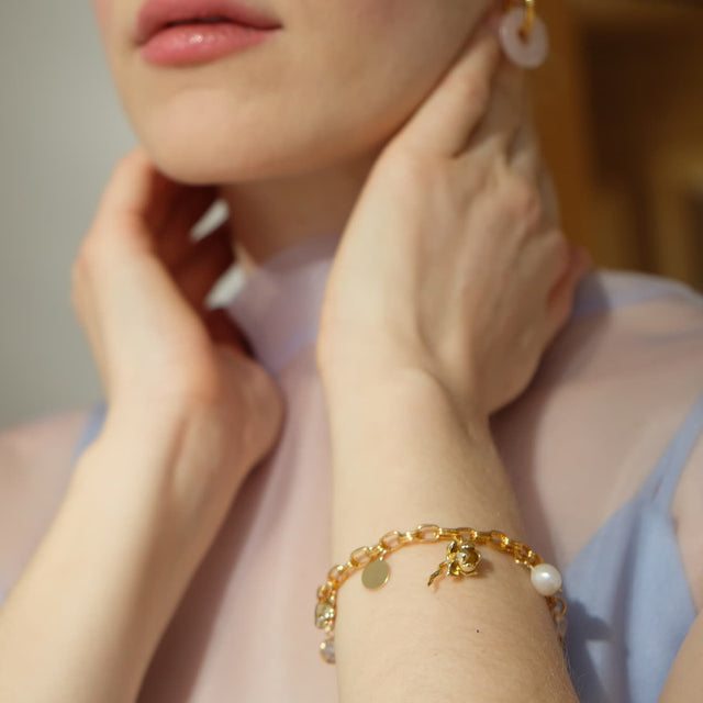 A light-skinned person touches their neck with both hands, wearing a transparent blue top. They sport a Noémiah gold bracelet—the Victoria Bracelet—featuring heart charms and hoop earrings, elegantly highlighting their jewelry. Focus is on hands and accessories.