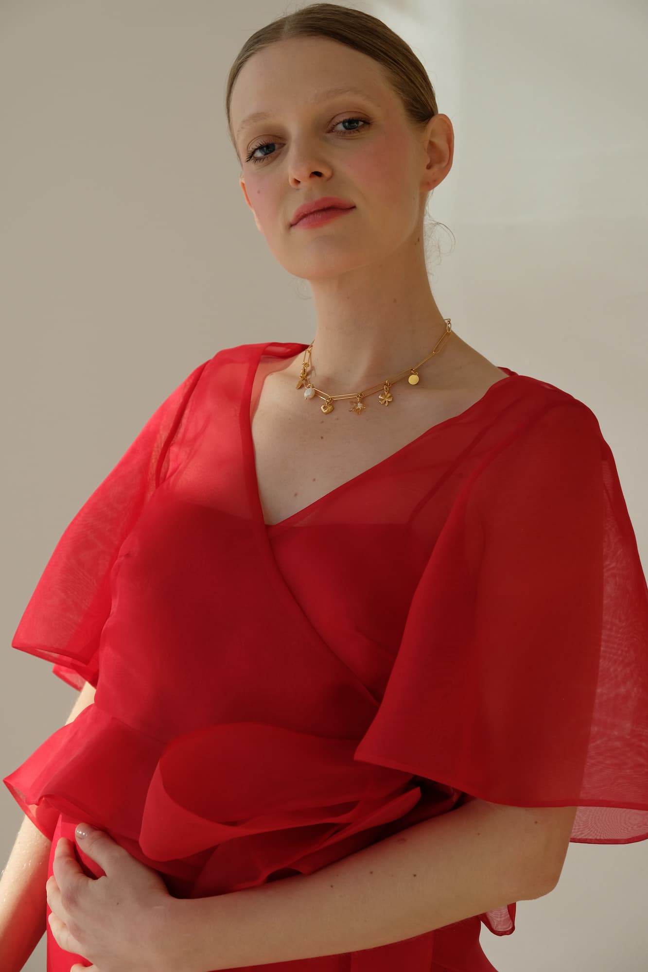 In a tranquil scene, a person in a red dress with fluttery sleeves and layered design poses serenely, wearing the Noémiah Violette Necklace adorned with an orange crystal heart. One hand rests on their abdomen as the softly lit background completes the ambiance.