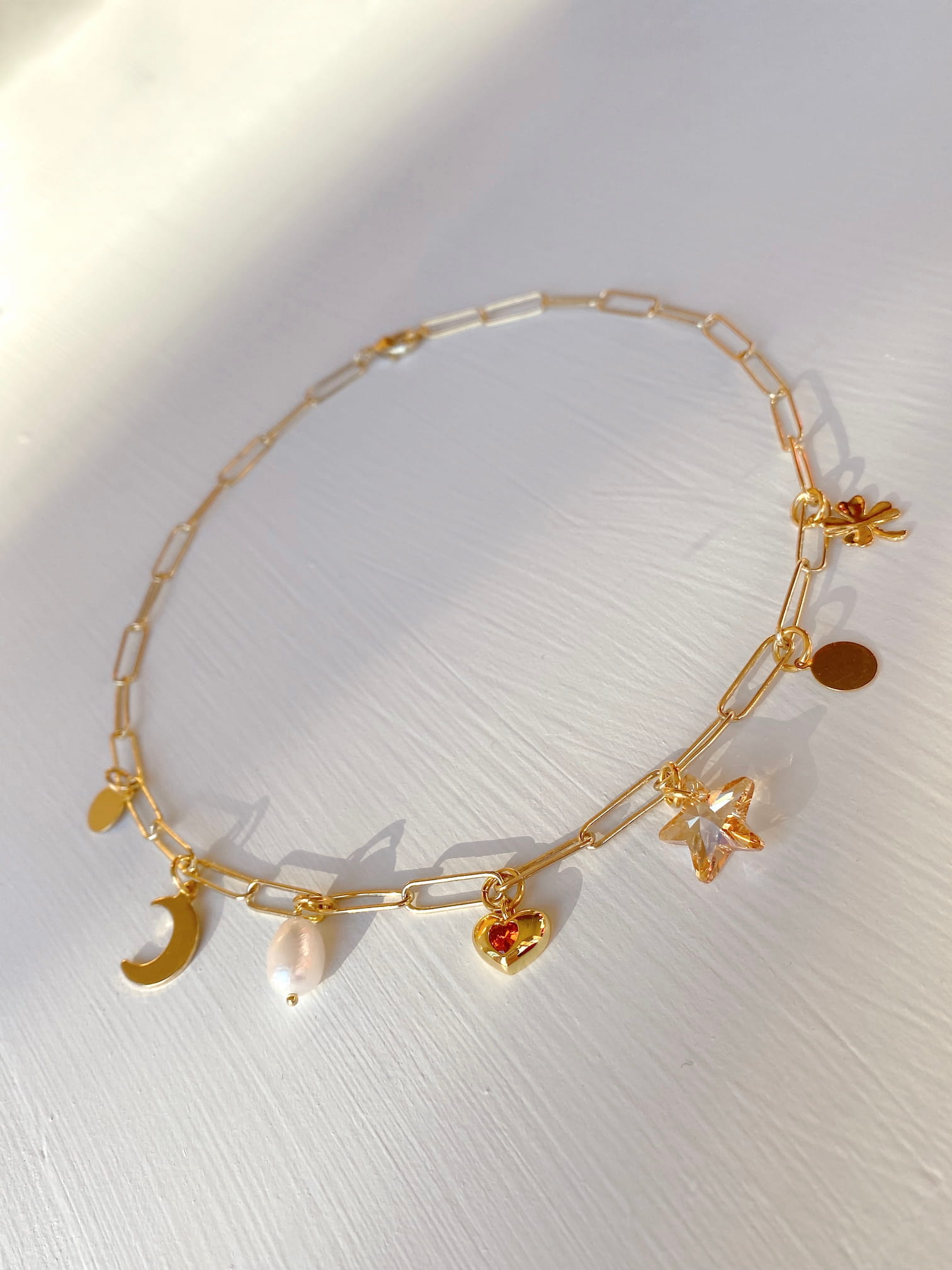 The Violette Necklace by Noémiah, featuring a gold chain adorned with a crescent moon, a freshwater natural pearl, and a Swarovski® star crystal charm, is displayed on a white surface. It glimmers with elegance in the soft, warm lighting.