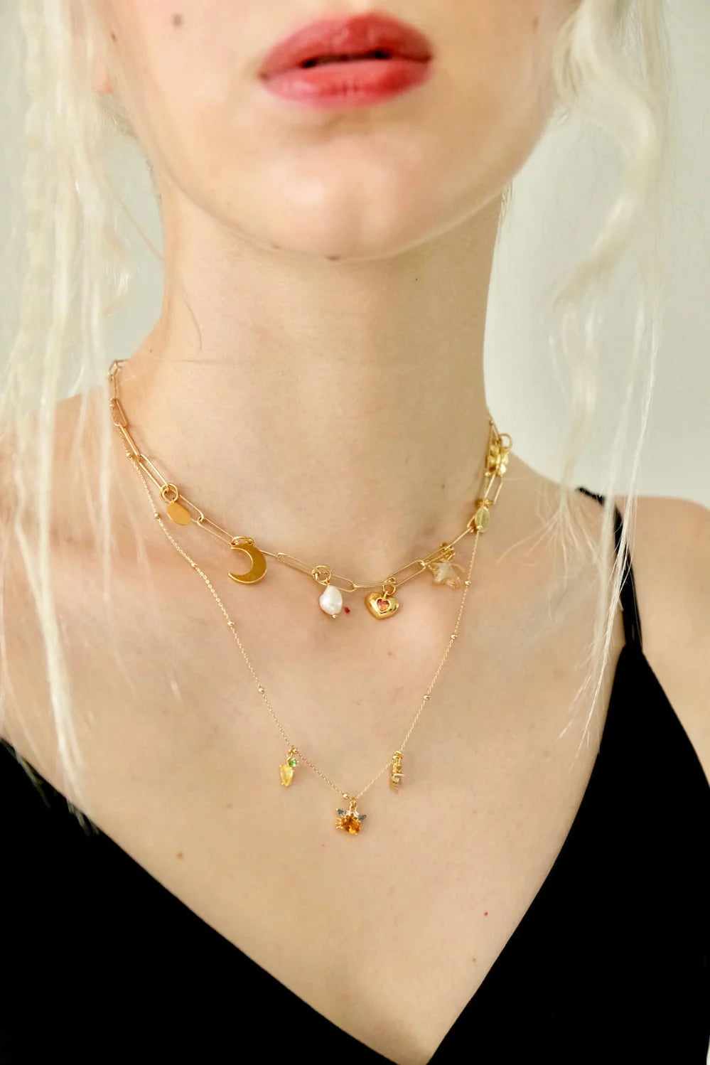 A person with light skin is dressed in a black top and layered gold Violette Necklaces by Noémiah. These necklaces, showcasing a Swarovski® star crystal alongside moon and colorful stone charms, beautifully enhance their slightly parted lips and light blonde hair.