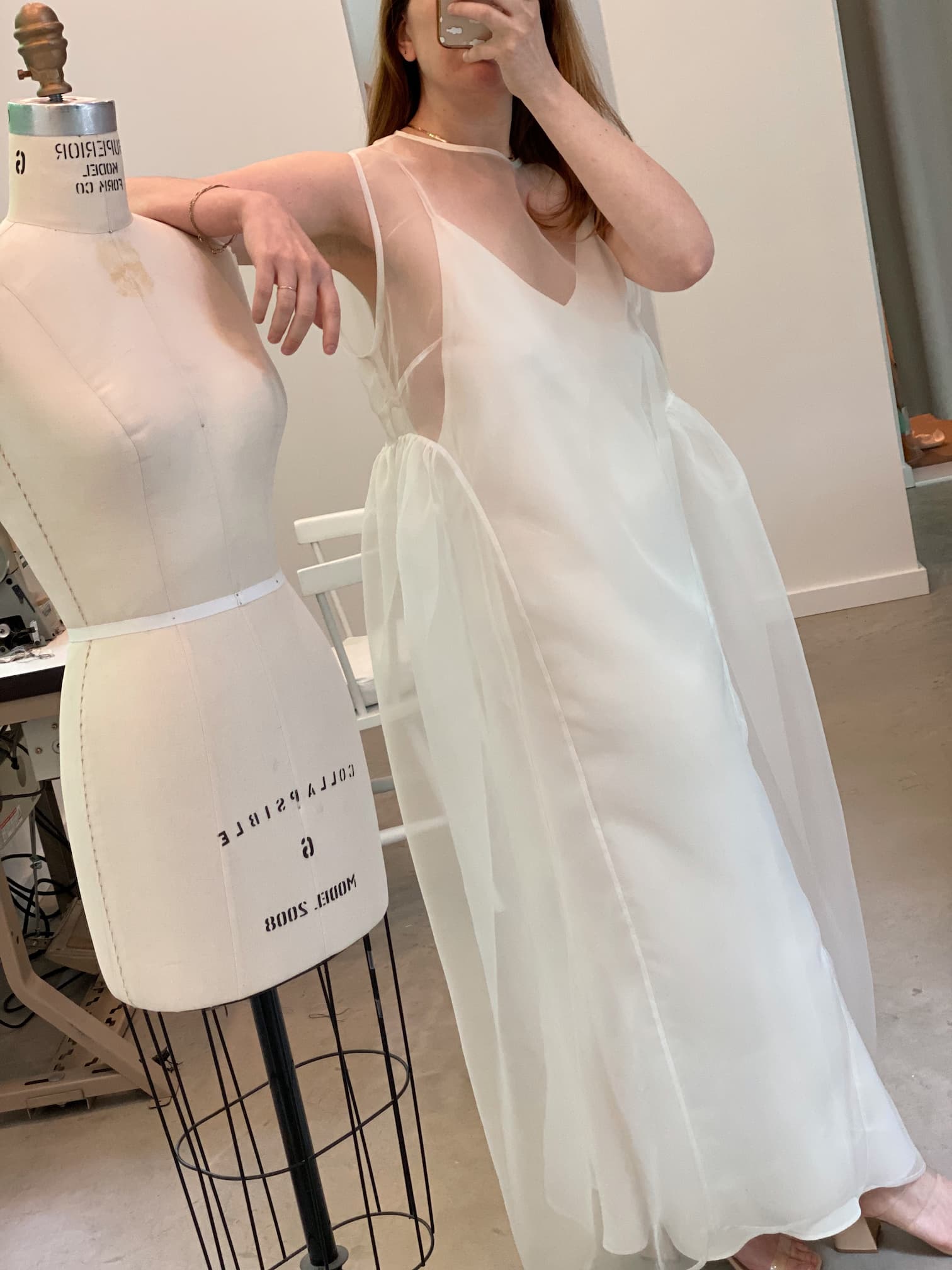 A person in a sleeveless, floor-length Noémiah Vivi Dress stands next to a dress form mannequin. The gown is adorned with an elegant cutout detail and features a flowing design with a sheer organza silk overlay. The room showcases a minimalist, modern interior with soft lighting.