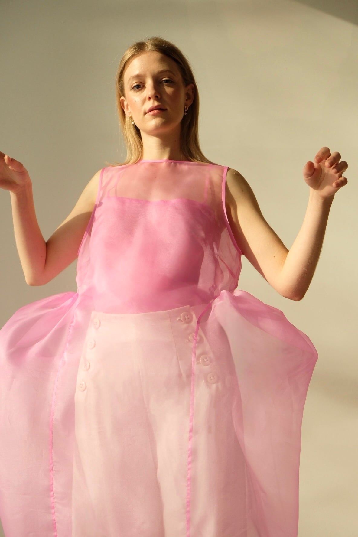 A person wearing a sheer, flowing Vivi Dress from Noémiah, featuring elegant cutout details, poses with arms slightly raised against a plain background. The lighting casts soft shadows, highlighting the texture and transparency of the organza silk fabric.