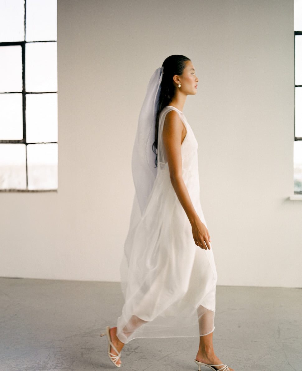 A woman wearing the Noémiah Vivi Dress, crafted from organza silk and featuring elegant cutout details, walks gracefully in a minimalist room with industrial-style windows, complemented by strappy heels. The soft natural light adds an ethereal quality to the scene.