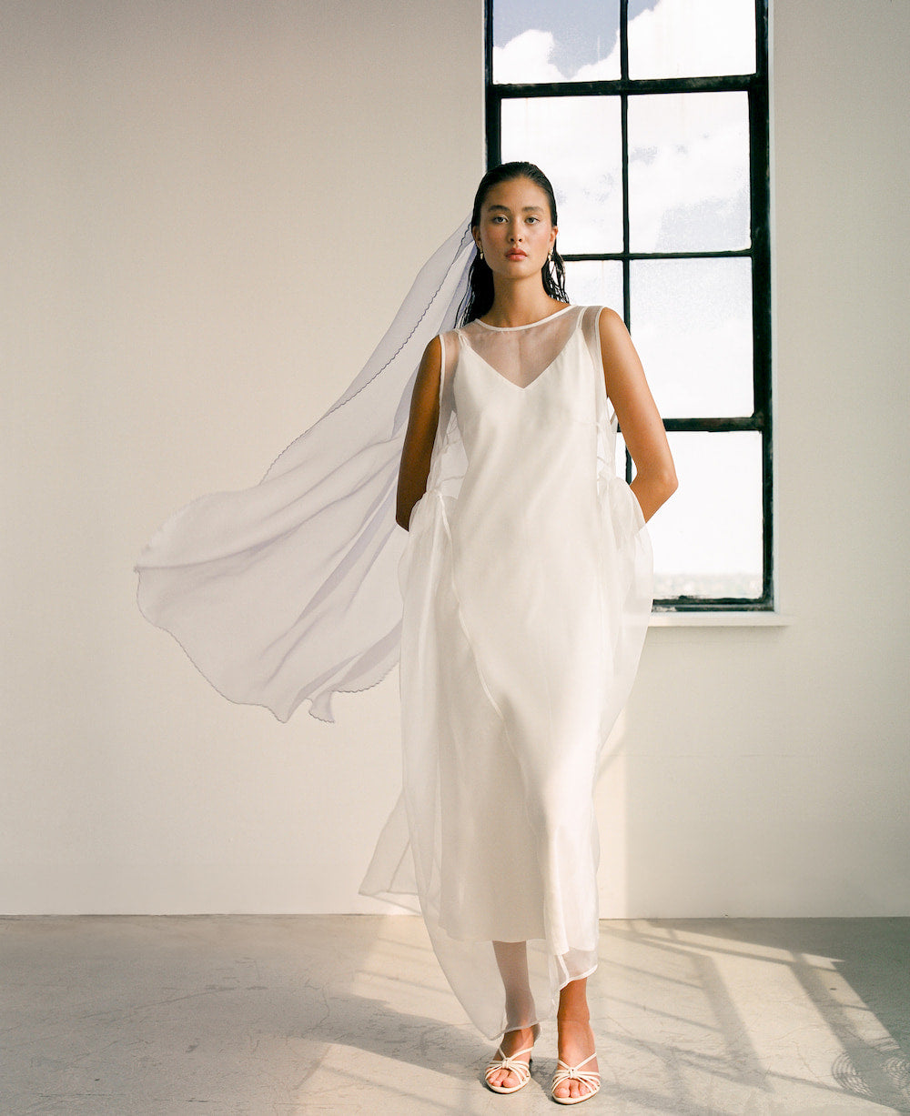 A woman stands in a sunlit room wearing the flowing Vivi Dress by Noémiah, made from organza silk and featuring an elegant cutout detail. She gazes forward with a serene expression while a large window behind her reveals a partly cloudy sky. She is standing on a light-colored floor.