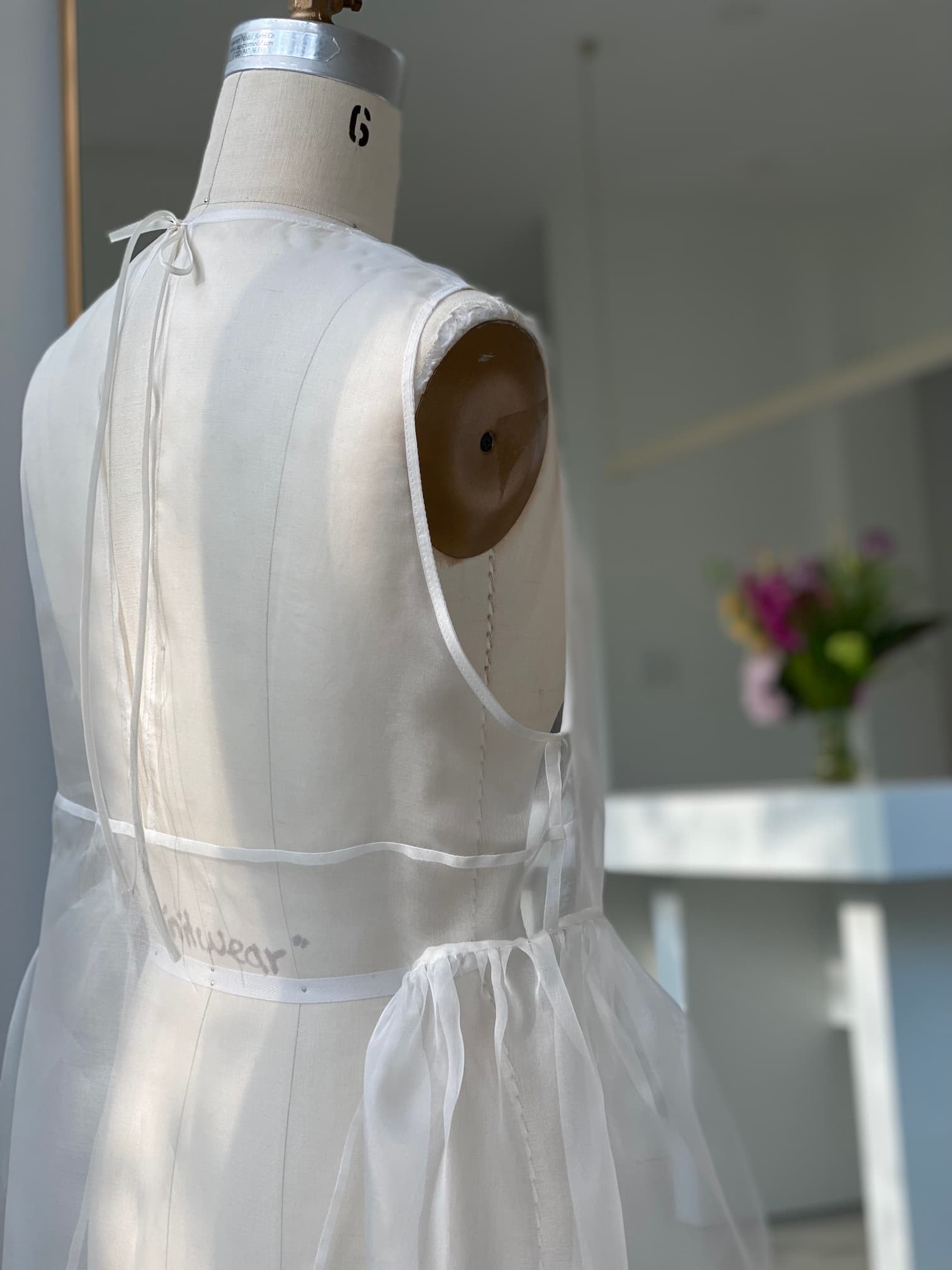 A dress form displays a sheer, white Noémiah Vivi Dress featuring sophisticated cutout details and delicate ties in a brightly lit room. In the background, there is a sleek white counter adorned with a vase filled with vibrant flowers.