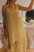 A person is indoors wearing the Noémiah Vivi Dress in flowing sheer yellow. The organza silk dress, characterized by its thin straps and elegant cutout details, falls gracefully. Visible tattoos decorate the arm, while a table in the background is adorned with yellow floral decorations.