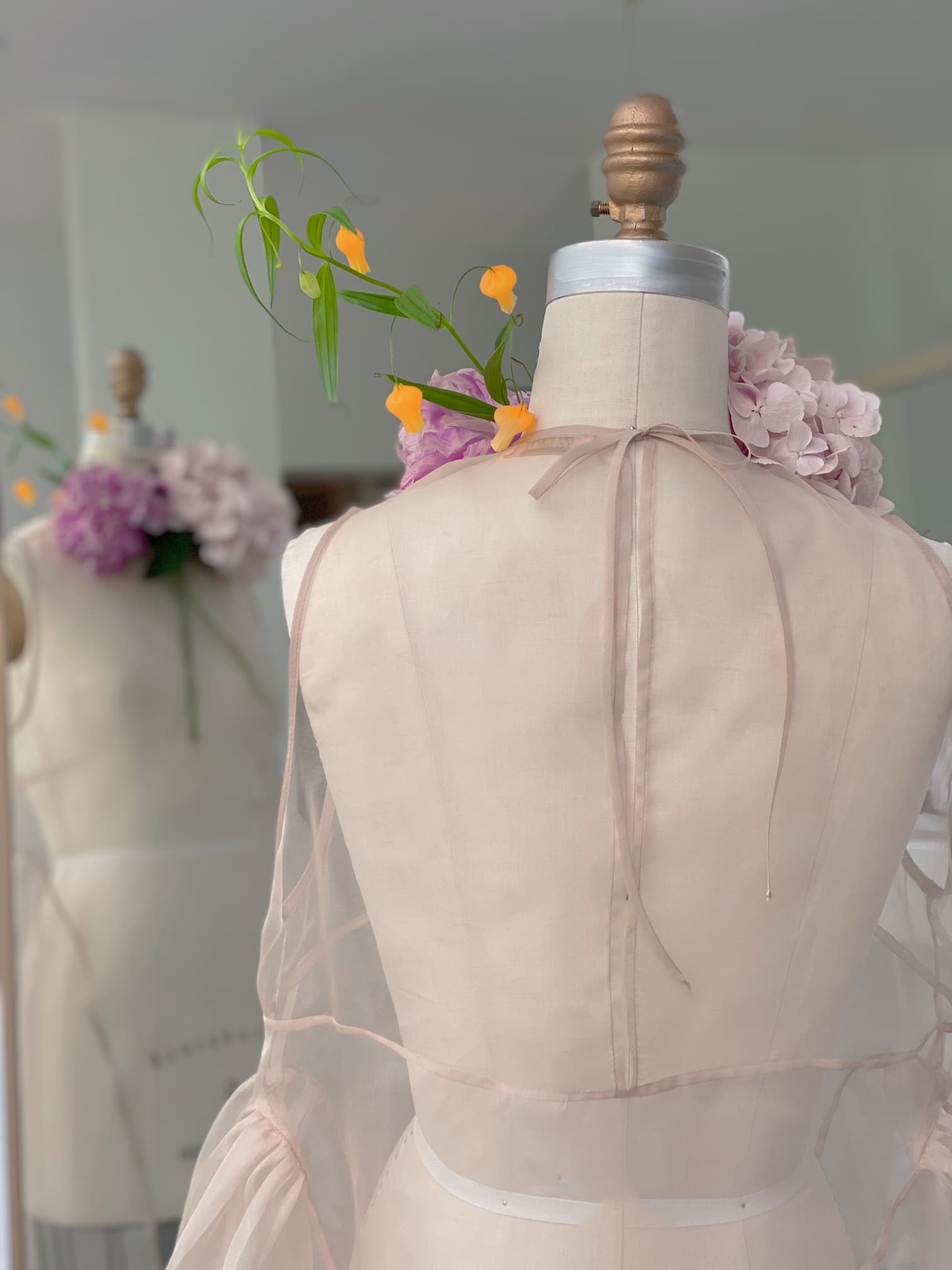 A dress form draped in sheer organza silk stands in a room, adorned with orange and purple flowers. Another dress form is partially visible in the background, reflecting a creative and elegant fashion design setting reminiscent of the Vivi Dress collection by Noémiah.