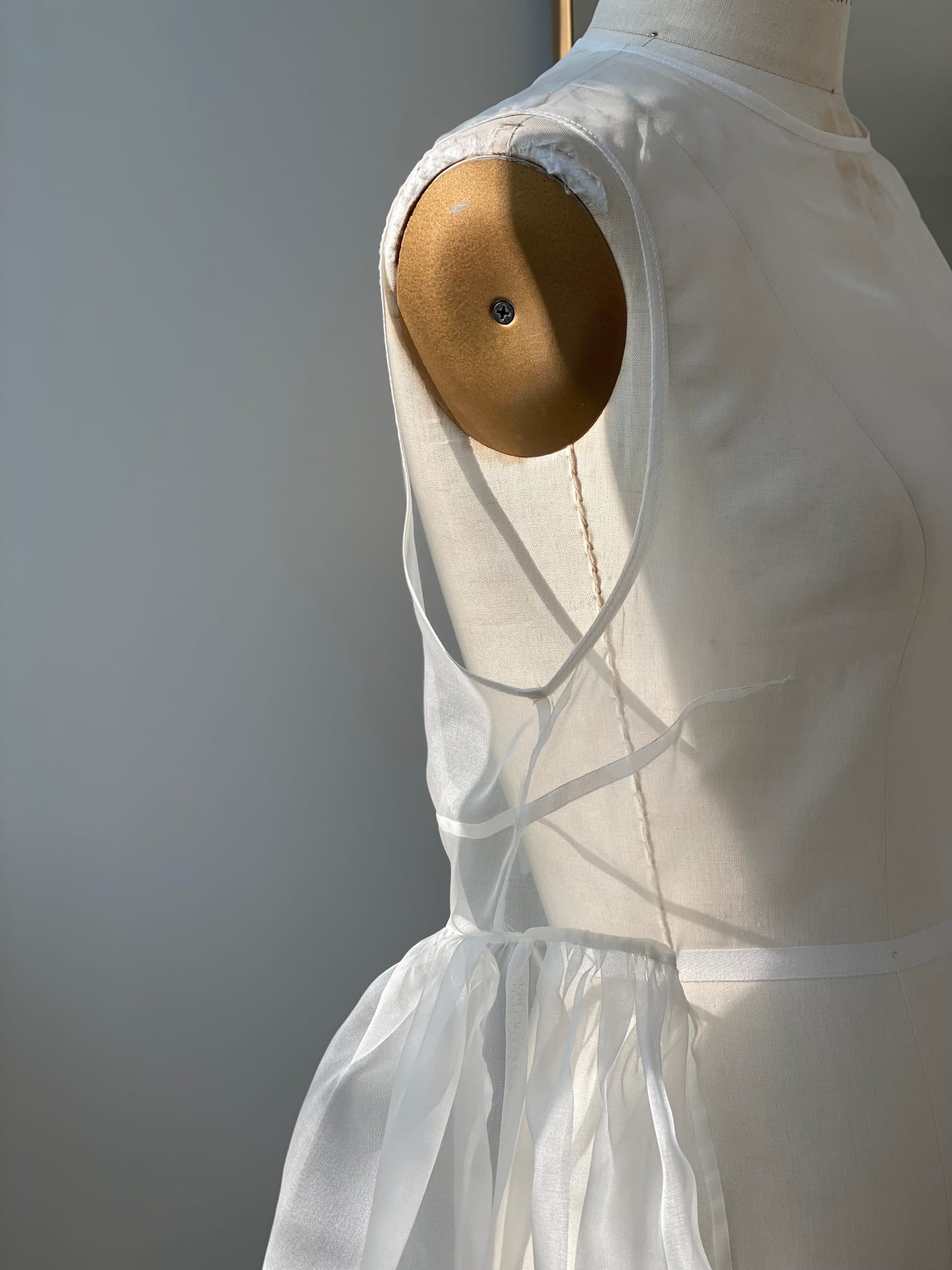 A close-up captures a dress form adorned with the Vivi Dress by Noémiah, its sleeveless design elegantly touched by sunlight. The dress showcases sheer organza silk details at the sleeve openings and a gathered waist, offering a soft and sophisticated aesthetic.