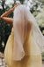 A person wearing a Noémiah Vivi Dress, characterized by its elegant cutout detail, stands with their back to the camera, holding a sheer white veil of organza silk above their head. They are outdoors on a balcony, surrounded by a softly blurred backdrop of trees.