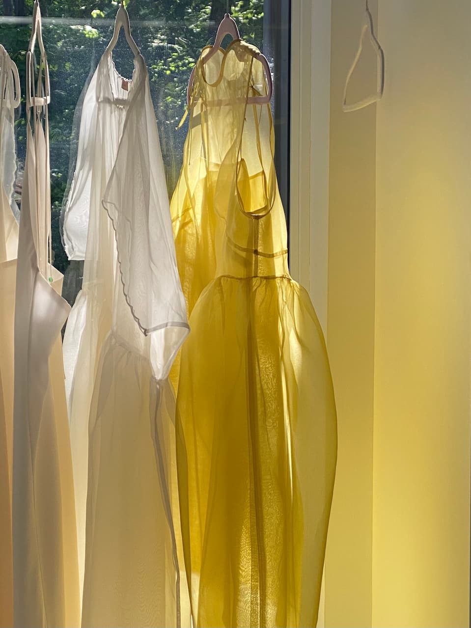 Two sheer dresses, one a white masterpiece with delicate trimming and the other the vibrant Vivi Dress by Noémiah crafted in organza silk, hang by a window in the soft sunlight. The natural light creates a gentle, airy atmosphere, accentuating the fabrics' elegant cutout details and translucency.