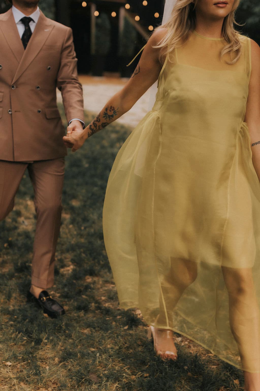 A person in a sheer yellow Noémiah Vivi dress holds hands with a person in a tan double-breasted suit while walking outdoors.