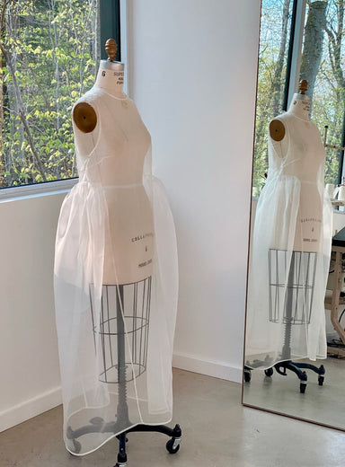 A mannequin adorned with the Noémiah Vivi Dress, made from sheer organza silk, is positioned near a large studio window. The dress features an elegantly mirrored cutout detail, complementing the view of leafy green trees outside and enhancing the room's minimalist and airy ambiance.