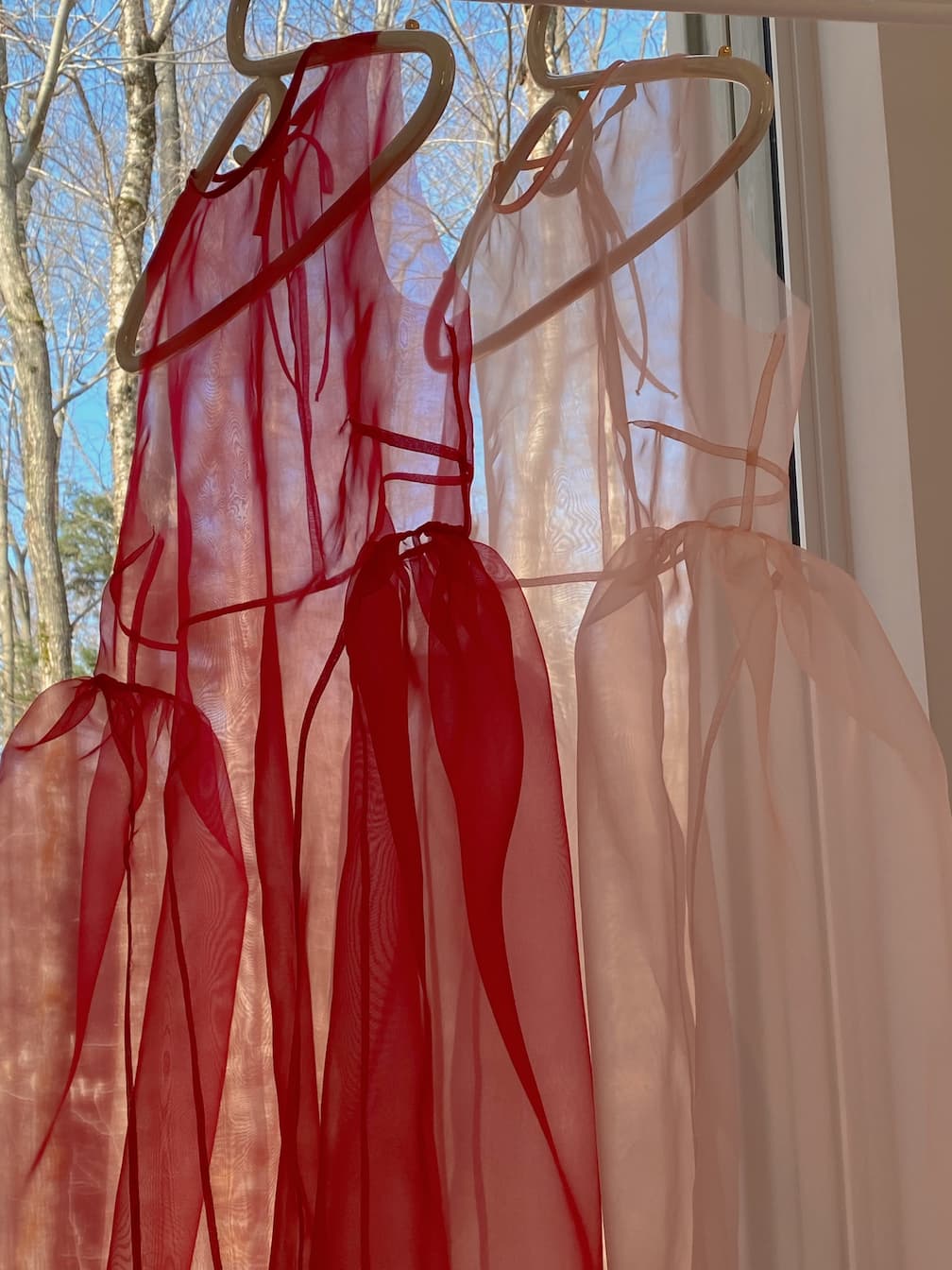 Sheer red and light pink Vivi dresses from Noémiah, crafted from organza silk, hang on a window with bare trees in the background. Sunlight filters through, creating a soft glow that highlights the elegant cutout details of each dress.