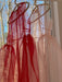 Sheer red and light pink Vivi dresses from Noémiah, crafted from organza silk, hang on a window with bare trees in the background. Sunlight filters through, creating a soft glow that highlights the elegant cutout details of each dress.