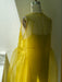 A mannequin adorned with a sheer, bright yellow Vivi Dress by Noémiah is positioned near a sunlit window. Sunlight streams through the organza silk material, accentuating its translucency and flowy texture. The dress boasts a neck tie, sophisticated cutout details, and layered, airy fabric.