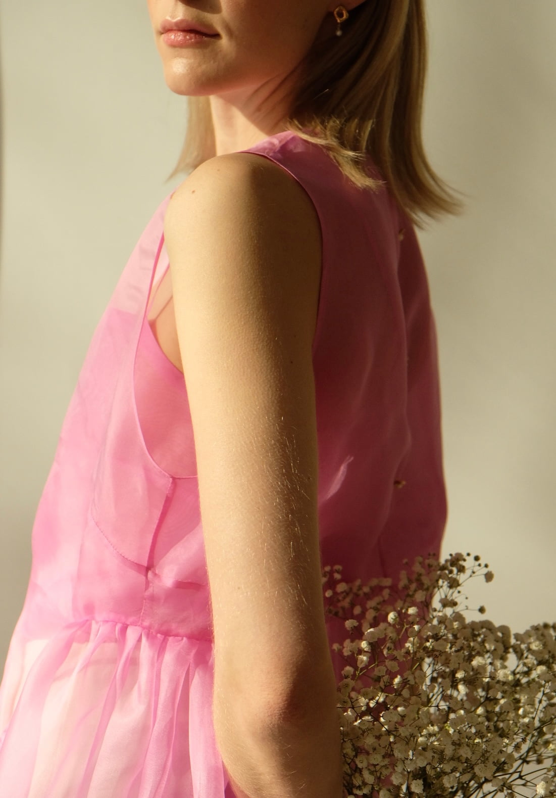 A person is captured in a close-up shot, donning the sophisticated Vivi Dress by Noémiah. They stand against a softly illuminated backdrop, gracefully holding a cluster of small white flowers. The image focuses on their upper body and arm, emphasizing the intricate cutout design of the pink organza silk fabric.