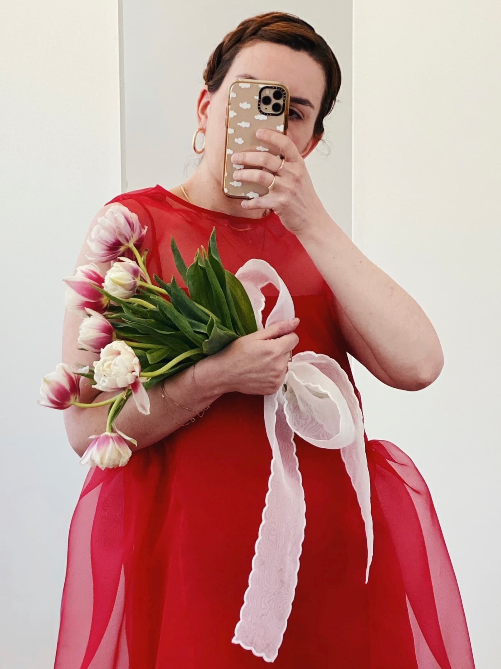 Clad in the elegant Vivi Dress by Noémiah with its striking red hue, a person takes a mirror selfie, highlighting the graceful cutout detail while holding a bouquet of pink tulips wrapped in white ribbon.