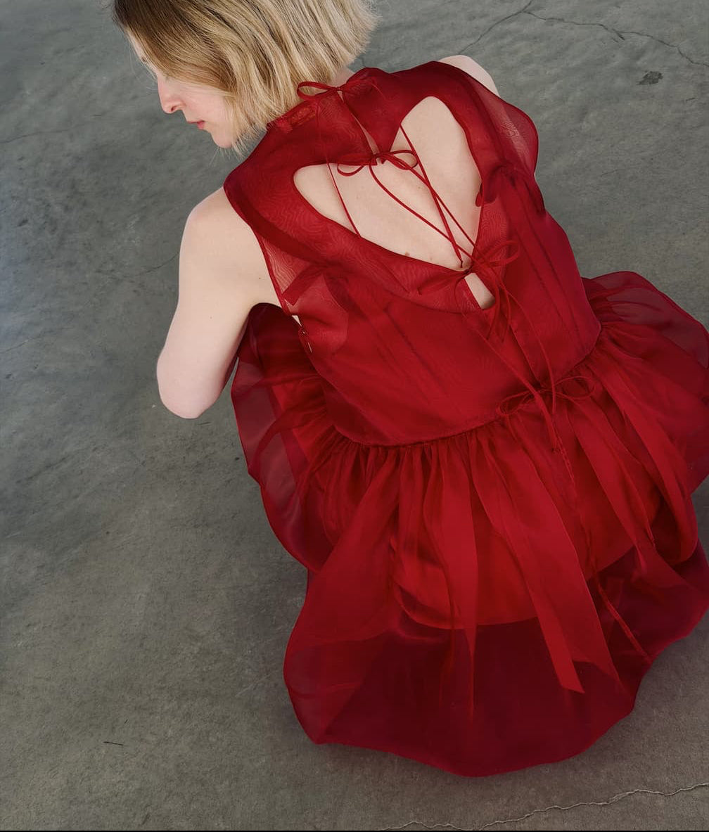 A person with short blonde hair sits on a gray concrete floor, wearing the Noémiah Viviane Dress—a layered red silk organza piece with ribbon details and an open back. The photo is shot from above and slightly behind.