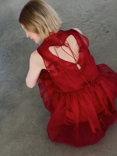 A person with short blonde hair sits on a gray concrete floor, wearing the Noémiah Viviane Dress—a layered red silk organza piece with ribbon details and an open back. The photo is shot from above and slightly behind.