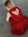 A person with short blonde hair sits on a gray concrete floor, wearing the Noémiah Viviane Dress—a layered red silk organza piece with ribbon details and an open back. The photo is shot from above and slightly behind.