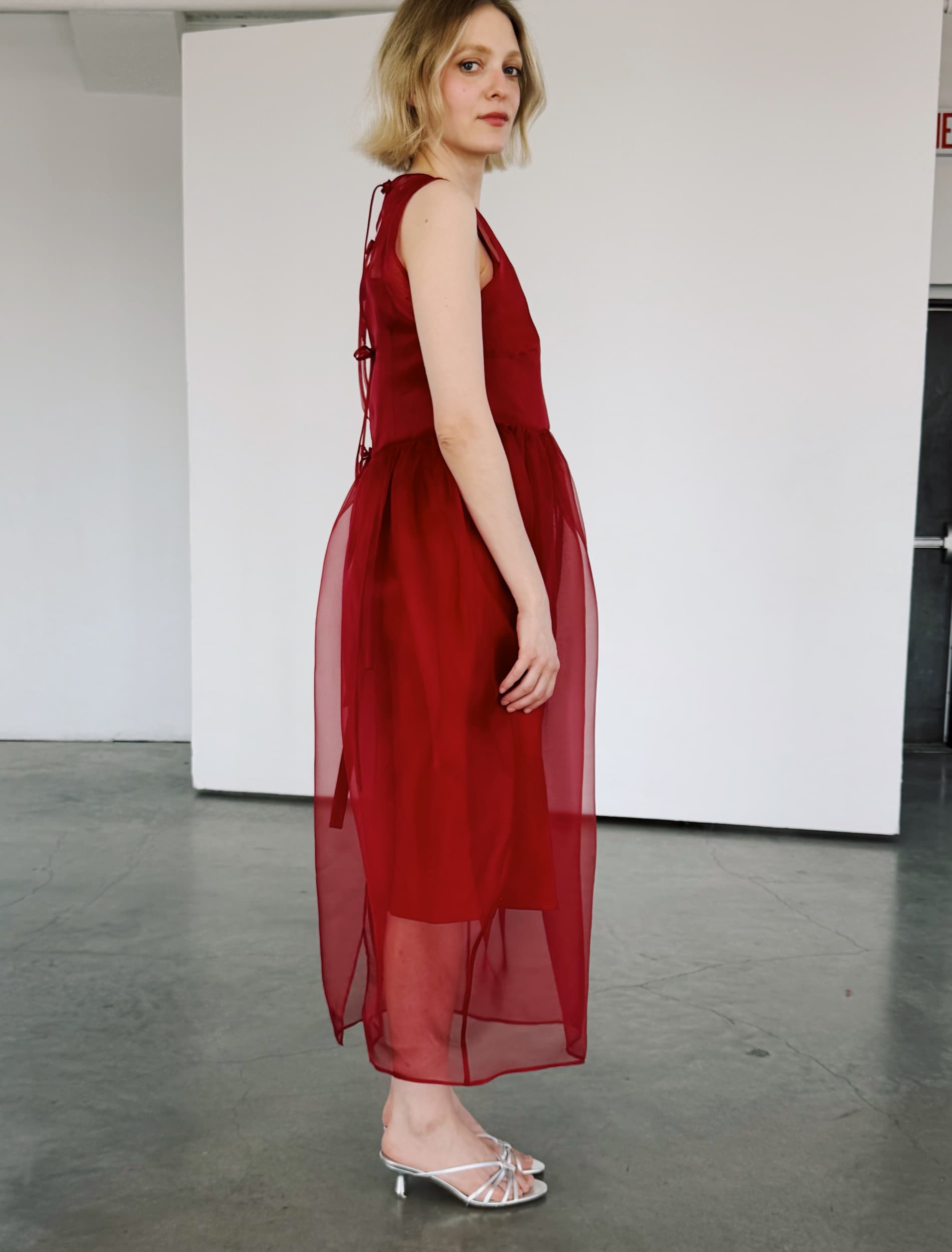 A blonde woman wears the Viviane Dress by Noémiah and silver open-toe heels, standing sideways in a minimalist room with concrete floors and white walls.
