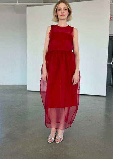 A woman stands indoors against a white wall on a concrete floor, wearing the Viviane Dress by Noémiah—a sleeveless, semi-sheer silk organza design with a fitted bodice and flowing skirt—paired with silver strappy heels.
