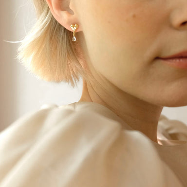 A close-up showcases a person with short blonde hair wearing a single Yuna earring by Noémiah, featuring an elegant teardrop design. They are dressed in a soft beige textured blouse, and the softly blurred background enhances the elegance of both the earring and attire.