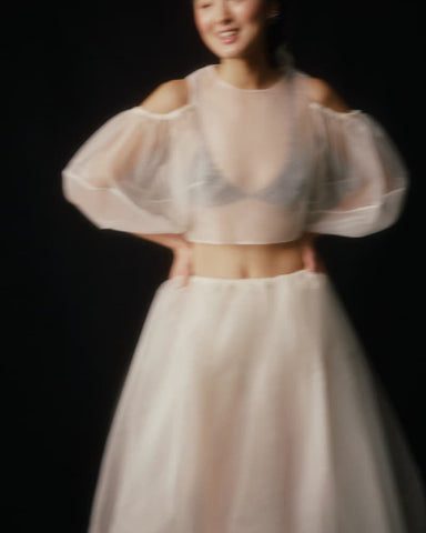 In a blurred image against a dark background, a person wearing the Zhou Skirt by Noémiah appears ethereal in a sheer top with puffy sleeves and a flowing silk organza midi skirt. Smiling with their hands on their hips, they evoke the dreamy charm reminiscent of a bridal ensemble.