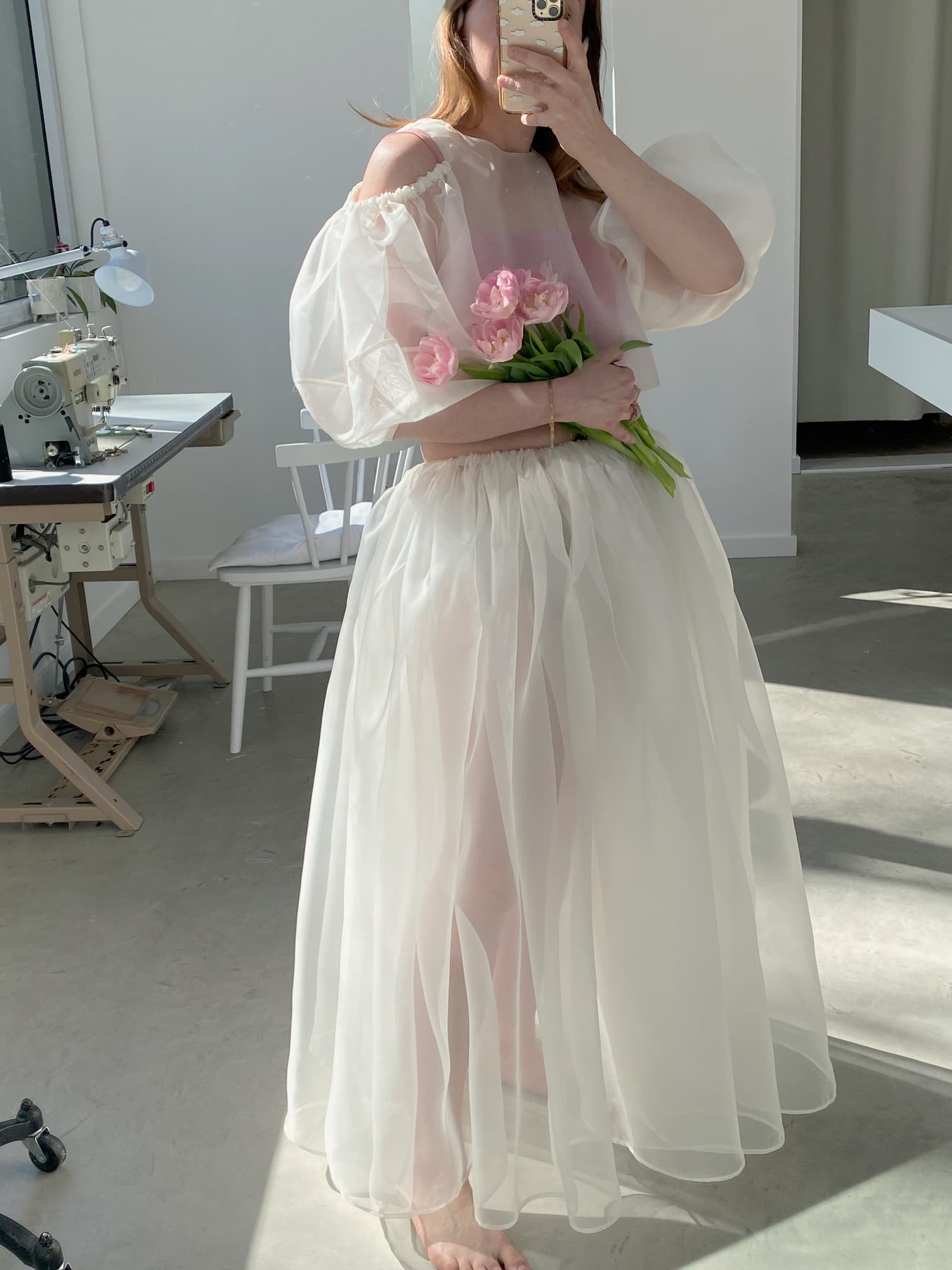 In a studio, someone is taking a selfie while holding a bunch of pink tulips and wearing the Zhou Skirt from Noémiah, a sheer white dress with puffed sleeves. The silk organza fabric beautifully catches the light, and in the background, there's a sewing machine suggesting creative bridal ensemble compositions.