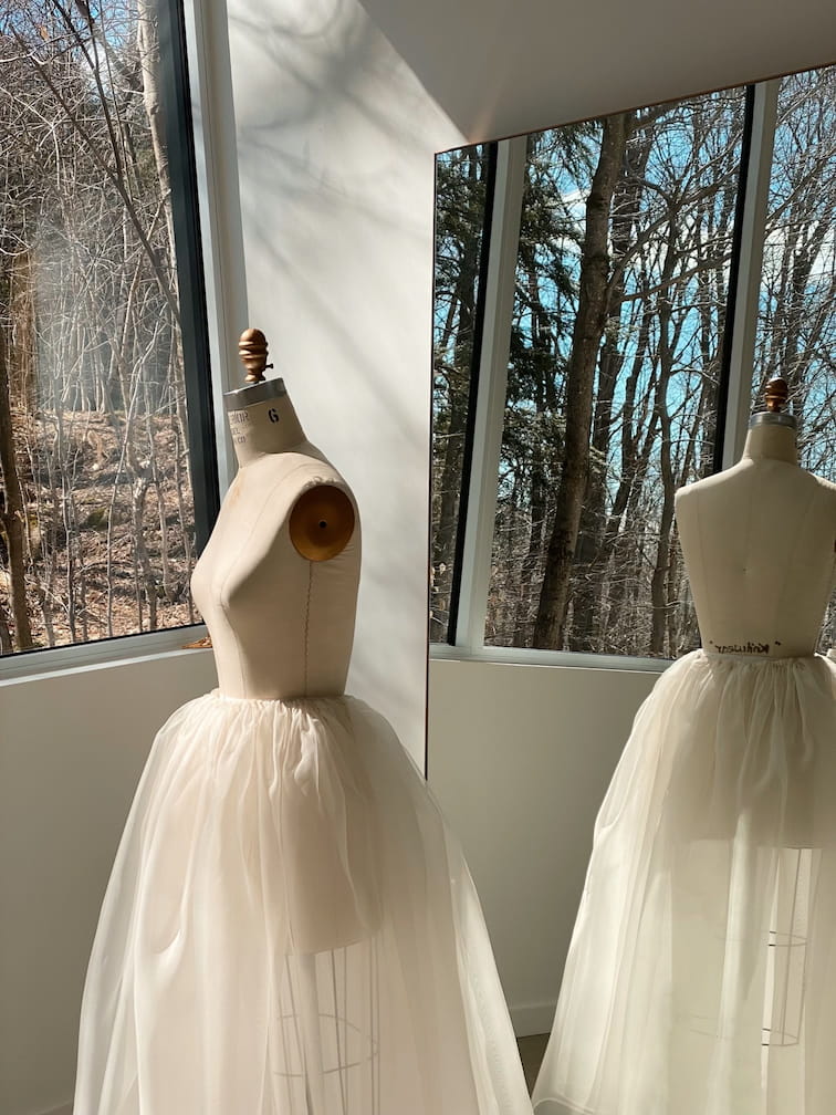 A Zhou Skirt from Noémiah stands by large windows, elegantly showcasing its light, airy silk organza material. Sunlight streams in from outside, where bare trees are visible, creating a serene and artistic atmosphere perfect for a bridal ensemble.