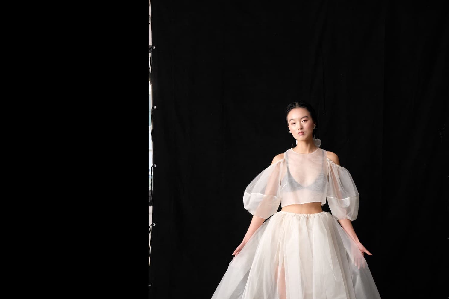 A person stands against a black background wearing the Zhou Skirt by Noémiah, crafted from flowing, semi-transparent silk organza with puffed sleeves. This layered bridal ensemble complements their serene pose as they look directly at the camera.