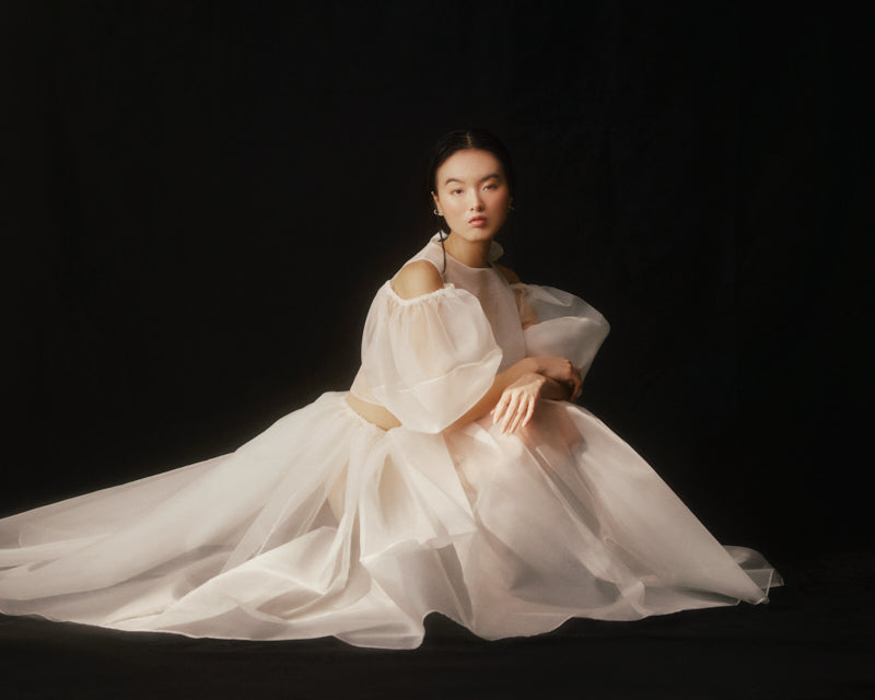 A person sits elegantly on a dark background, wearing the Zhou Skirt by Noémiah—a flowing, sheer white design with puffed sleeves. The silk organza fabric of the skirt drapes softly around them, creating a serene and ethereal appearance.