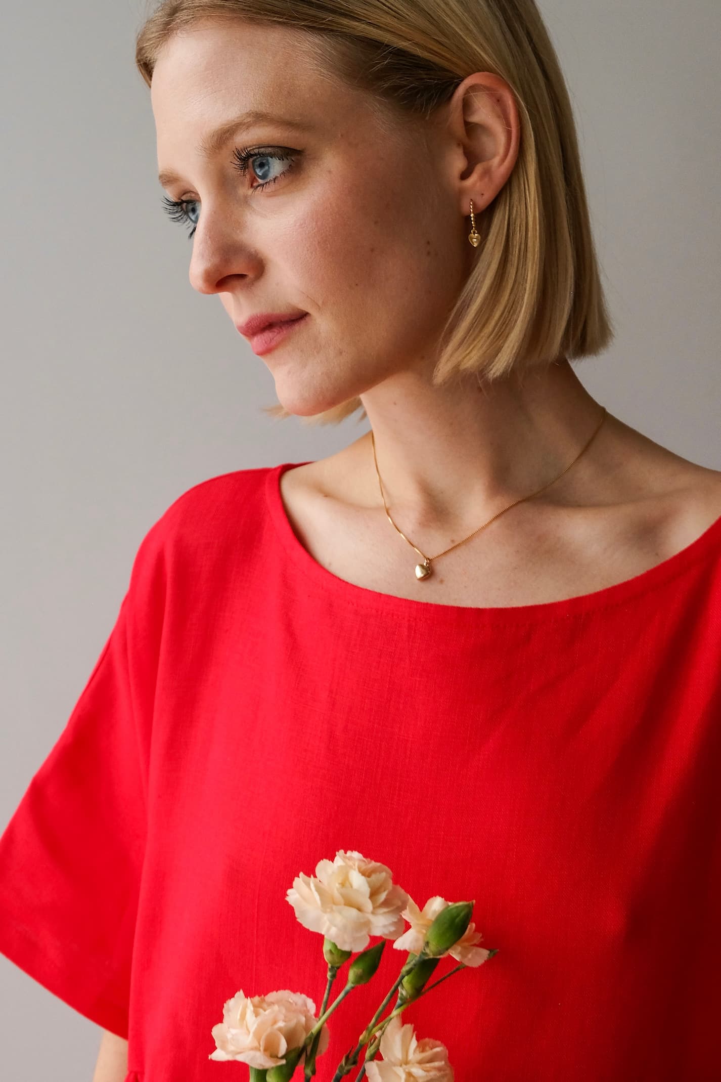 A person with short blonde hair, wearing a red top and a 14K gold filled necklace featuring the Noémiah Zoé Pendant, holds a small bouquet of light pink flowers against a plain, neutral background.