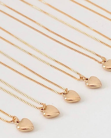 Close-up of several Noémiah Zoé Pendant necklaces laid out on a flat surface, each featuring a small, shiny heart charm and crafted from 14K gold filled chain. The necklaces are uniformly arranged and aligned, creating a visually pleasing, symmetrical pattern reminiscent of their elegant style.