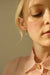 A close-up of a person wearing a pale pink blouse and Noémiah's Alaska Earrings, featuring gold-plated hoops with a delicate charm, partially visible against a neutral background. The person appears pensive as sunlight illuminates their face.