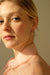 A person with blond hair is wearing a light pink blouse and the Alaska Earrings by Noémiah. They are looking towards the camera with a neutral expression against a softly lit beige background.