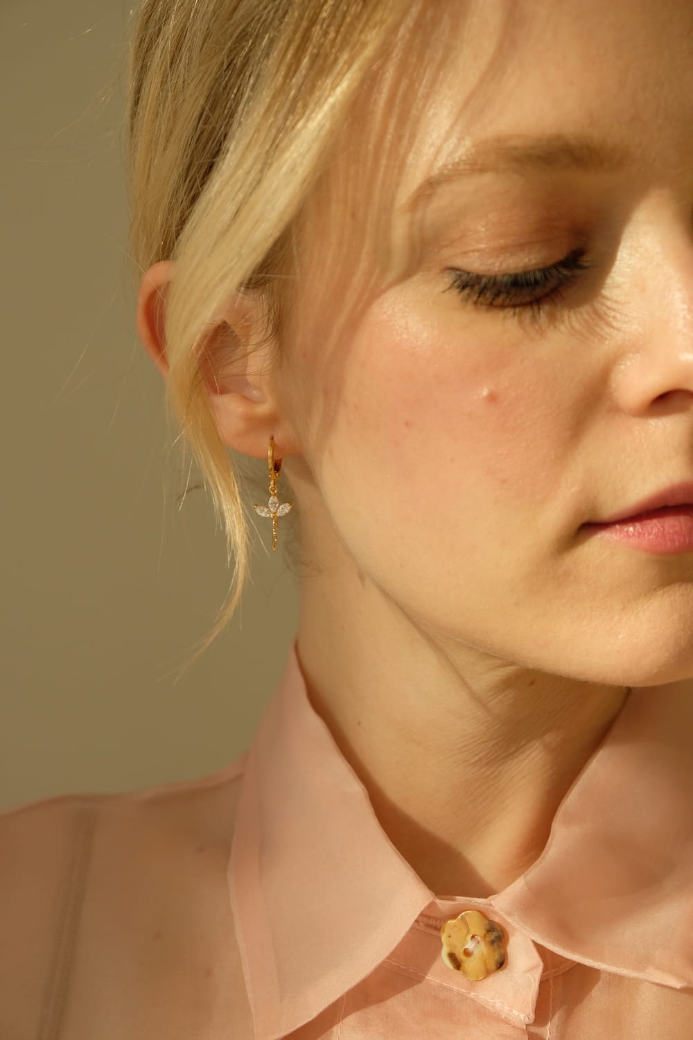 A close-up of a woman wearing a light pink blouse with a cat button. She has light hair and is adorned with the elegant Alaska Earrings by Noémiah, which feature a sophisticated gold-plated design with a small drop detail. The image captures half of her serene face against a soft background, highlighting her delicate jewelry.