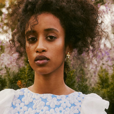 A woman with curly hair stands outdoors in front of blooming flowers. She wears a blue and white floral-patterned top and the Noémiah Annette Earrings adorned with freshwater pearls, gazing calmly at the camera. The tarnish-resistant gold earrings perfectly complement the soft-focus background featuring greens and purples.