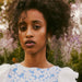 A woman with curly hair stands outdoors in front of blooming flowers. She wears a blue and white floral-patterned top and the Noémiah Annette Earrings adorned with freshwater pearls, gazing calmly at the camera. The tarnish-resistant gold earrings perfectly complement the soft-focus background featuring greens and purples.