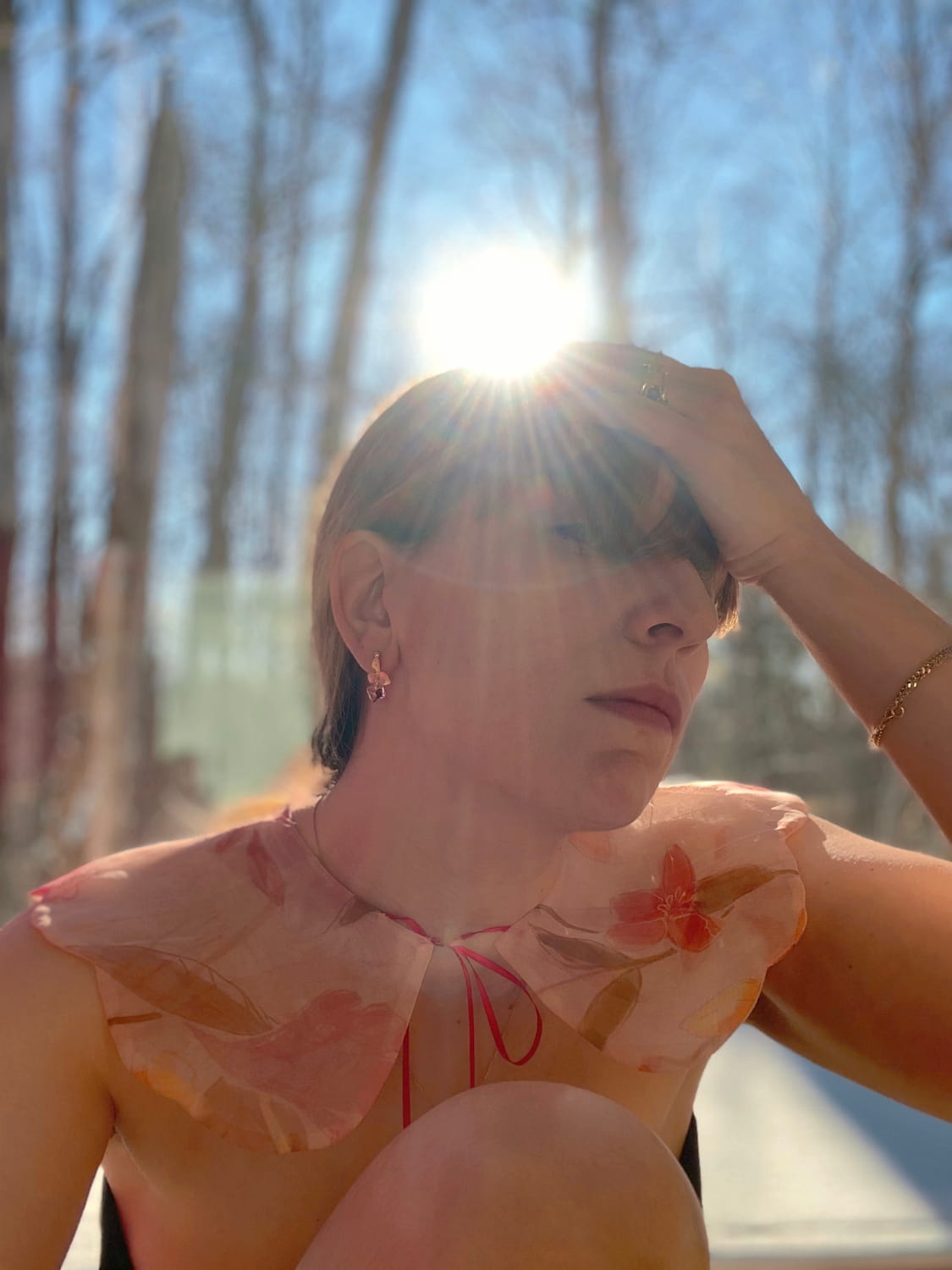 Sunlight streams in as a person with short hair sits by a window, adorned in a sheer, floral collar and wearing Noémiah's tarnish-resistant Annette Earrings featuring Swarovski crystals. With their hand resting on their forehead, the earrings catch the light against the backdrop of trees outside.