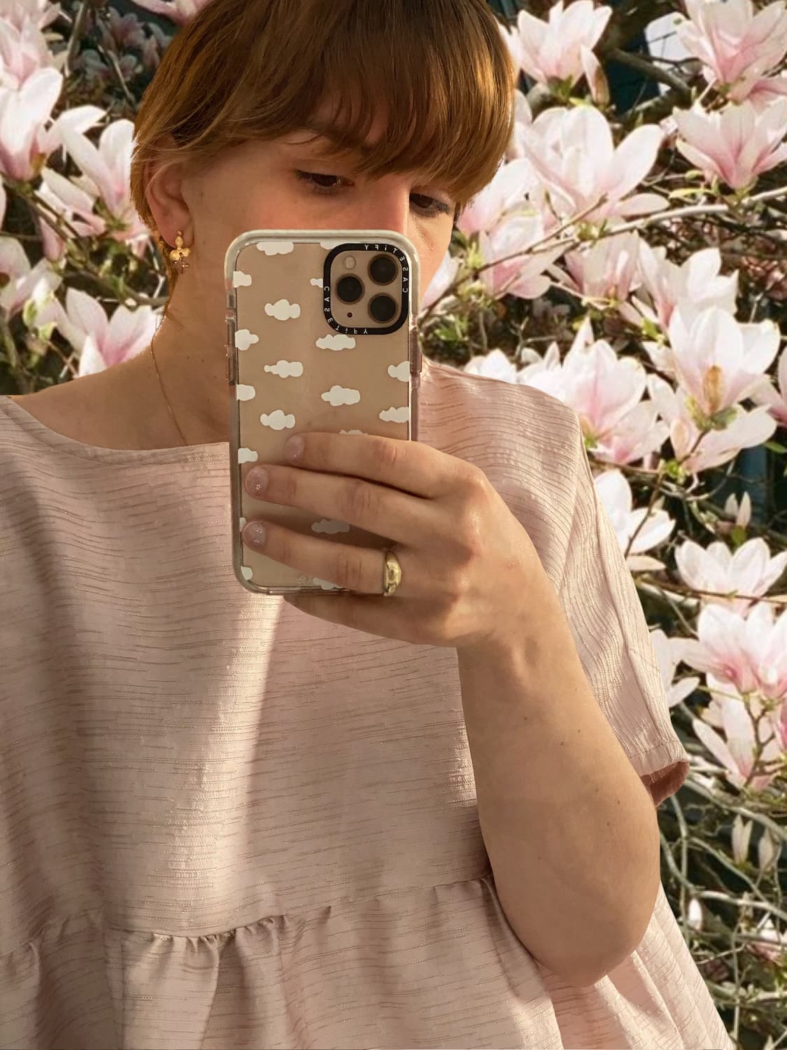 A person captures a mirror selfie with a phone, donned in a textured pink top that glistens with accents from Noémiah's Annette Earrings embellished with Swarovski crystals. Blooming pink magnolia flowers enhance the elegance of the scene in the background.