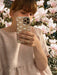 A person captures a mirror selfie with a phone, donned in a textured pink top that glistens with accents from Noémiah's Annette Earrings embellished with Swarovski crystals. Blooming pink magnolia flowers enhance the elegance of the scene in the background.