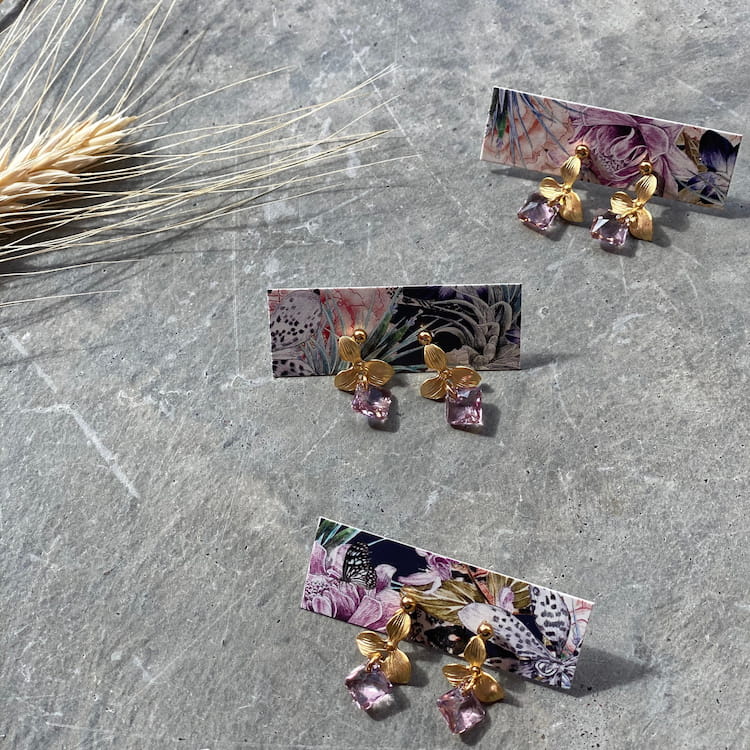 Three pairs of Noémiah's Annette Earrings — Swarovski, featuring gold flower designs with pink gemstone drops, shimmer gracefully on floral-patterned cards atop a textured concrete surface. Tarnish-resistant and ideal for daily wear, these exquisite earrings are accompanied by a small bundle of wheat to their left.