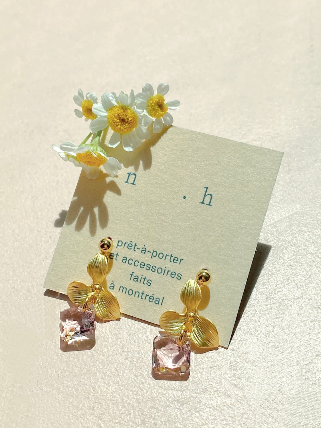 The Annette Earrings — Swarovski by Noémiah, featuring gold floral designs with pink Swarovski crystals, are elegantly presented on a card. This card contains French text about fashion and accessories from Montreal. These tarnish-resistant earrings harmonize beautifully with the small white and yellow daisies positioned above on a light-colored surface.