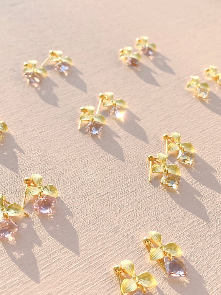 The Annette Earrings by Noémiah, featuring Swarovski crystals set in gold and shaped like flowers, are arranged diagonally on a textured pale pink surface, casting soft shadows from the sunlight. Ideal for everyday wear, these earrings provide elegance and are tarnish resistant.